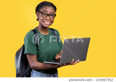 Happy Black Student Girl Using Laptop Computer Posing Smiling To Camera Standing Over Yellow Background. Studio Shot. E-Learning And Online Education. Educational Gadgets Concept Happy Black Student Girl Using Laptop Computer Posing Smiling To Camera Standing Over Yellow Background. Studio Shot. E-Learning And Online Education. Educational Gadgets Concept 134067562