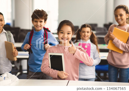 Education And Technology Concept. Smiling small asian girl holding digital tablet and showing thumbs up sign gesture. Diverse group of international classmates standing with backpacks in background 134067565