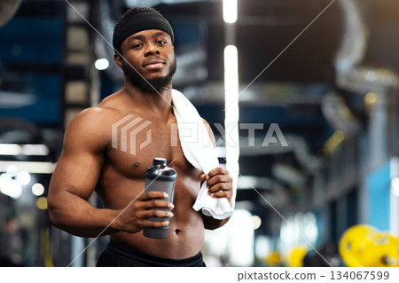 Rest after workout. Shirtless african american bodybuilder holding water or protein drink and towel at gym, empty space. Handsome black muscular man having break while exercising at modern gym Rest after workout. Shirtless african american bodybuilder holding water or protein drink and towel at gym, empty space. Handsome black muscular man having break while exercising at modern gym 134067599