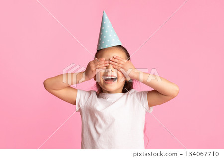 Having Fun. Excited asian girl covering her eyes, wearing party hat isolated over pastel pink studio wall, free space Having Fun. Excited asian girl covering her eyes, wearing party hat isolated over pastel pink studio wall, free space 134067710