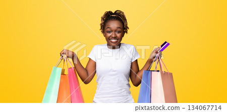 Happy black lady with shopping bags and credit card smiling over yellow studio background, empty space. African american woman shopaholic holding bunch of purchases and unlimited credit card 134067714