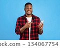 African Guy Pointing Finger At Smartphone Standing Over Blue Studio Background. Great Mobile Application Concept African Guy Pointing Finger At Smartphone Standing Over Blue Studio Background. Great Mobile Application Concept 134067754