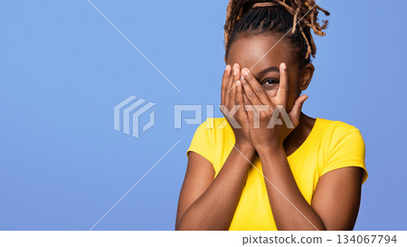 Cute young black woman closing her face with palms, looking with one eye through her fingers at camera, purple studio background, empty space. Cheerful african american lady playing hide and seek 134067794