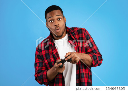 Black Millennial Guy Playing Video Games Holding Gamepad Standing Over Blue Studio Background. Gaming Concept Black Millennial Guy Playing Video Games Holding Gamepad Standing Over Blue Studio Background. Gaming Concept 134067805