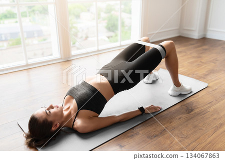 Fitness Concept. Athletic woman in black sportwear training with elastic resistance band above knees in fitness studio or in living room. Lady doing glute bridge exercise for buttocks, legs and core 134067863