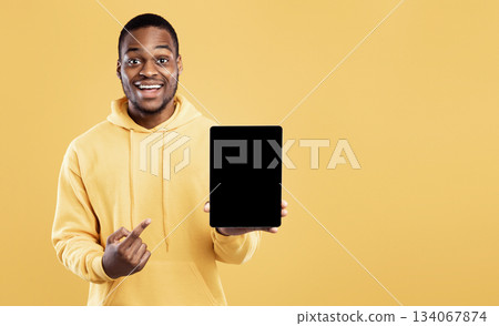 Black Man Pointing At Digital Tablet Empty Screen Holding Computer Posing Over Yellow Studio Background, Smiling To Camera. Mockup For Great Website Or Applicatioin Advertisement 134067874