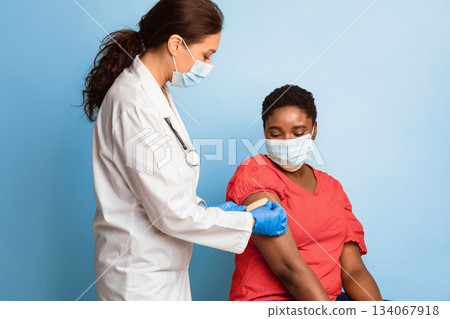 Antiviral Immunization, Covid-19 Vaccination. Black Female Getting Vaccinated, Doctor Sticking Adhesive Bandage After Vaccine Injection On Patient's Arm Over Blue Background. Studio Shot 134067918
