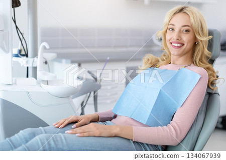 Side view of pretty blonde young woman patient sitting in dental chair and smiling, copy space. Cheerful millennial lady showing beautiful white smile after treatment at modern dental clinic Side view of pretty blonde young woman patient sitting in dental chair and smiling, copy space. Cheerful millennial lady showing beautiful white smile after treatment at modern dental clinic 134067939