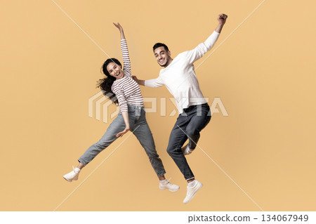 Playful Mood. Full Length Shot Of Happy Middle Eastern Couple Jumping In Air Over Yellow Studio Background, Cheerful Excited Arab Man And Woman Having Fun And Fooling Together, Copy Space Playful Mood. Full Length Shot Of Happy Middle Eastern Couple Jumping In Air Over Yellow Studio Background, Cheerful Excited Arab Man And Woman Having Fun And Fooling Together, Copy Space 134067949