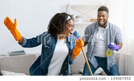 Happy Housekeeping. Cheerful African Couple Having Fun While Tidying Home, Woman Singing To Mop Handle, Making Concert In Living Room Done Happy Housekeeping. Cheerful African Couple Having Fun While Tidying Home, Woman Singing To Mop Handle, Making Concert In Living Room Done 134067958