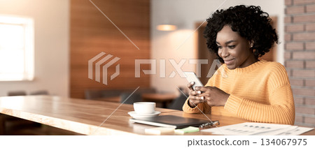 Busy cheerful black girl chatting at cafe, using modern smartphone and drinking tea, copy space Busy cheerful black girl chatting at cafe, using modern smartphone and drinking tea, copy space 134067975