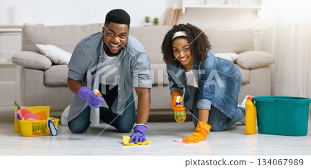 Household Chores Concept. Smiling black couple washing floor in their apartment with detergent spray and rag, making spring cleaning together Household Chores Concept. Smiling black couple washing floor in their apartment with detergent spray and rag, making spring cleaning together 134067989