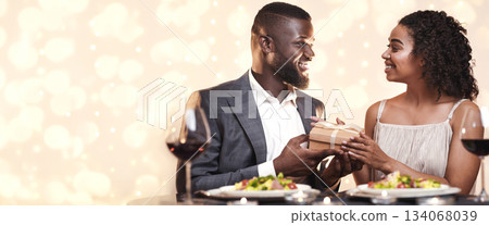 Valentine's present. Afro man giving his surprised girlfriend box of present. Couple sitting and drinking wine at restaurant Valentine's present. Afro man giving his surprised girlfriend box of present. Couple sitting and drinking wine at restaurant 134068039