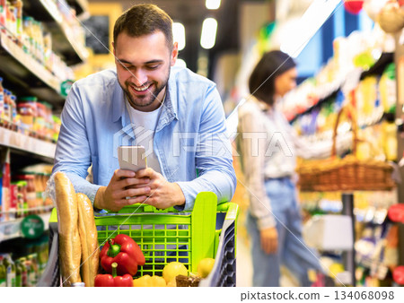 Smiling Male Customer Doing Grocery Shopping Using Smartphone Walking With Cart In Supermarket. Selective Focus. Man Using Groceries Shopping Application On Phone Bying Food In Super Market Smiling Male Customer Doing Grocery Shopping Using Smartphone Walking With Cart In Supermarket. Selective Focus. Man Using Groceries Shopping Application On Phone Bying Food In Super Market 134068098