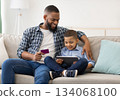 Online Shopping. Black Father And Son Using Digital Tablet Buying Things And Making Payment Via Credit Card Sitting On Couch At Home. Modern Family Shopping Concept Online Shopping. Black Father And Son Using Digital Tablet Buying Things And Making Payment Via Credit Card Sitting On Couch At Home. Modern Family Shopping Concept 134068100