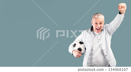 Celebrating Goal. Cheering senior man posing with soccer ball, making winner gesture, blue studio wall Celebrating Goal. Cheering senior man posing with soccer ball, making winner gesture, blue studio wall 134068107
