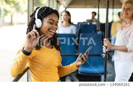 Portrait of cheerful young black woman passenger standing and dancing in bus near window using cell listening to music in wireless headphones, enjoying song having fun at public city transport 134068161