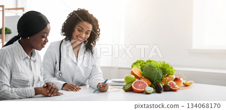 Cheerful african woman nutritionist making treatment plan for female patient, clinic interior 134068170