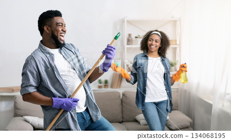 Tidying With Fun. Joyful Black Couple Fooling While Cleaning Flat Together, Man Using Mop Handle As Guitar And Laughing Tidying With Fun. Joyful Black Couple Fooling While Cleaning Flat Together, Man Using Mop Handle As Guitar And Laughing 134068195