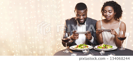Young black couple sitting in restaurant and taking pictures of food with mobile phone, food bloggers celebrating Valentine's Day 134068244