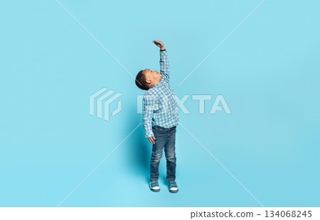 Little african american boy wishes he were taller, raising his hand to show how tall he wants to grow, child measuring his height, posing isolated on blue background, panorama, full length 134068245