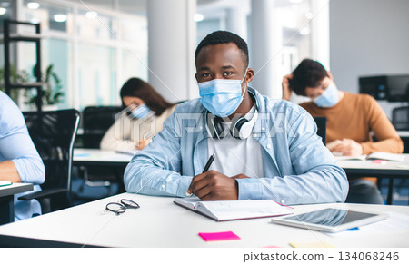 Modern Education During Pandemic Concept. Portrait of african american male student sitting at desk in classroom at university, wearing protective medical mask, writing in notebook, looking at camera 134068246