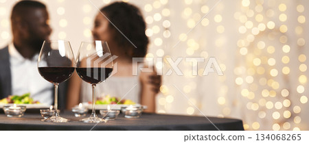 Selective focus on two glasses of wine over black couple in love, having festive dinner at restaurant Selective focus on two glasses of wine over black couple in love, having festive dinner at restaurant 134068265