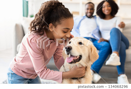 Portrait of smiling African American girl whispering something in the ear to her pet, telling secret and emracing happy dog, spending time together with best friend in living room, selective focus 134068296