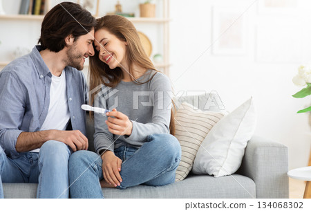 Happy family concept. Married couple bonding on couch and holding positive pregnancy test, home interior, free space 134068302