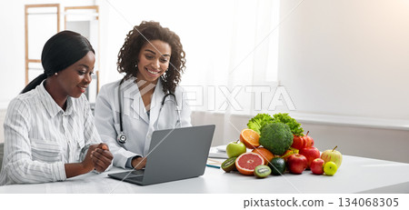 Useful medical service online. Smiling black woman nutritionist showing female patient information on laptop, clinic interior, copy space 134068305