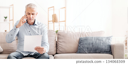 Discussing Document. Serious mature man holding paper and talking on phone, sitting on couch at home Discussing Document. Serious mature man holding paper and talking on phone, sitting on couch at home 134068309