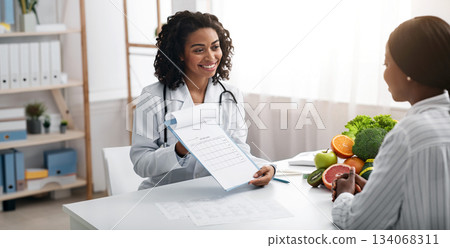 Cheerful african woman dietician showing female patient medical chart, clinic interior, empty space Cheerful african woman dietician showing female patient medical chart, clinic interior, empty space 134068311