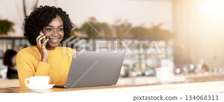 Smiling black businesswoman talking with client by phone while working with laptop at cafe, drinking coffee Smiling black businesswoman talking with client by phone while working with laptop at cafe, drinking coffee 134068313