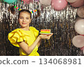 Happy girl in yellow holiday blouse with funny ponytails hairstyle and piece of cake in rainbow colors on festive background looks at sparklers 134068981