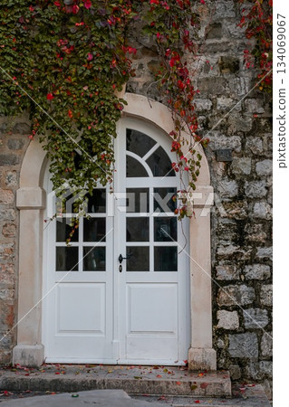Close-up photo of a white semi-circular door with glass set in a textured stone wall in the historic Old Town of Budva, Montenegro. Autumn leaves surround the doorway, highlighting the architectural Close-up photo of a white semi-circular door with glass set in a textured stone wall in the historic Old Town of Budva, Montenegro. Autumn leaves surround the doorway, highlighting the architectural 134069067