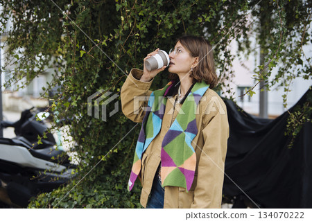 Woman with vibrant scarf and jacket drinks coffee near bike Woman with vibrant scarf and jacket drinks coffee near bike 134070222