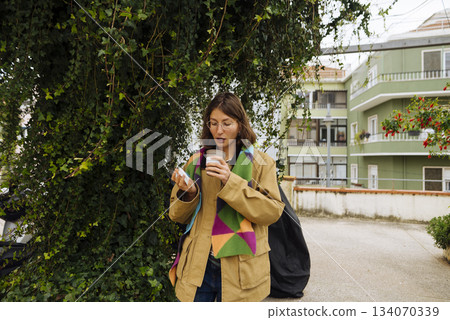 Young woman with backpack drinking coffee outdoors smiling warmly Young woman with backpack drinking coffee outdoors smiling warmly 134070339