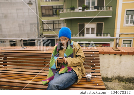 Woman in blue beanie on city bench with device and beverage Woman in blue beanie on city bench with device and beverage 134070352