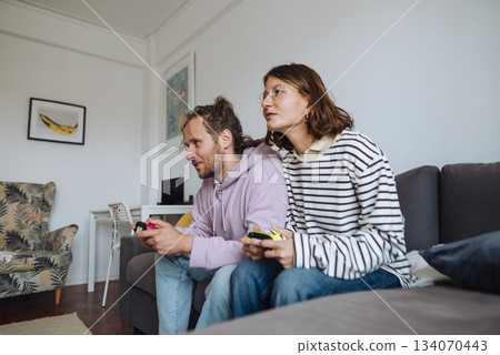 Diverse couple engages with controllers in cozy living space Diverse couple engages with controllers in cozy living space 134070443