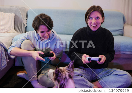 Two women sit on the floor playing a video game while their dog watches closely. The moment captures friendly competition and a cozy, relaxed home mood Two women sit on the floor playing a video game while their dog watches closely. The moment captures friendly competition and a cozy, relaxed home mood 134070959
