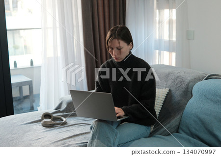 A woman works on a laptop while sitting on a sofa in a bright room, concentrating on tasks in a calm home environment A woman works on a laptop while sitting on a sofa in a bright room, concentrating on tasks in a calm home environment 134070997