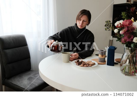 A woman pours tea into a mug while sitting at a round table with snacks beside her. The scene captures a peaceful break and a warm daily ritual 134071002
