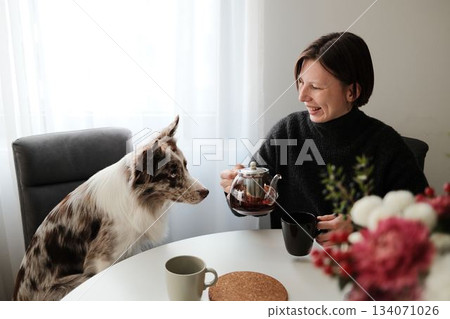 A woman laughs while holding a teapot as her dog curiously watches, capturing a warm and playful domestic interaction. The scene shows companionship and relaxed morning mood 134071026