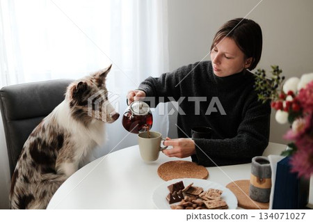 A woman pours tea at a breakfast table while her dog watches attentively, creating a warm domestic moment. The scene reflects companionship, calm routine, and quiet morning atmosphere A woman pours tea at a breakfast table while her dog watches attentively, creating a warm domestic moment. The scene reflects companionship, calm routine, and quiet morning atmosphere 134071027