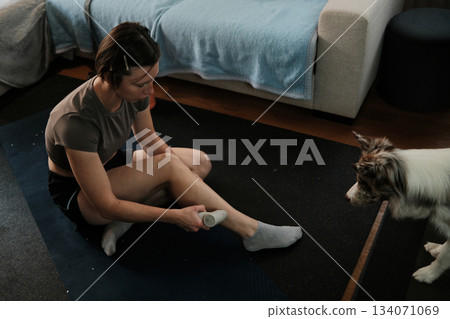 A woman uses a massage device on her leg while the border collie watches attentively. The moment conveys care, calmness, and domestic balance A woman uses a massage device on her leg while the border collie watches attentively. The moment conveys care, calmness, and domestic balance 134071069