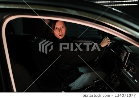 A woman drives a car out of a dim underground garage, illuminated by soft artificial light. The scene reflects daily commuting, independence, and modern mobility. 134071077