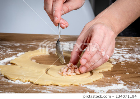 Hands Preparing Dough With Spoonful of Meat Filling on Floured Wooden Surface 134071194