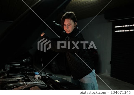 A woman opens the car hood and checks the engine compartment in an underground garage. The moment shows routine vehicle inspection and responsible car ownership 134071345