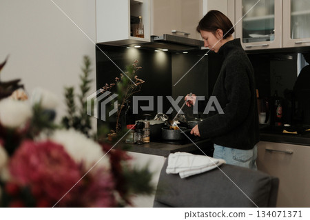 A woman cooks in a cozy kitchen, stirring food in a pot under soft lights and creating a warm, calm everyday atmosphere 134071371