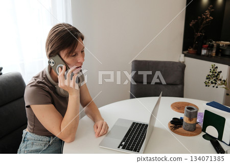 A woman talks on her phone while working at her laptop, combining digital and verbal communication in a home setting 134071385